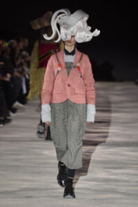 Runway model in pink plaid blazer with floral embroidery over gray check pants and a large white sculptural hat.