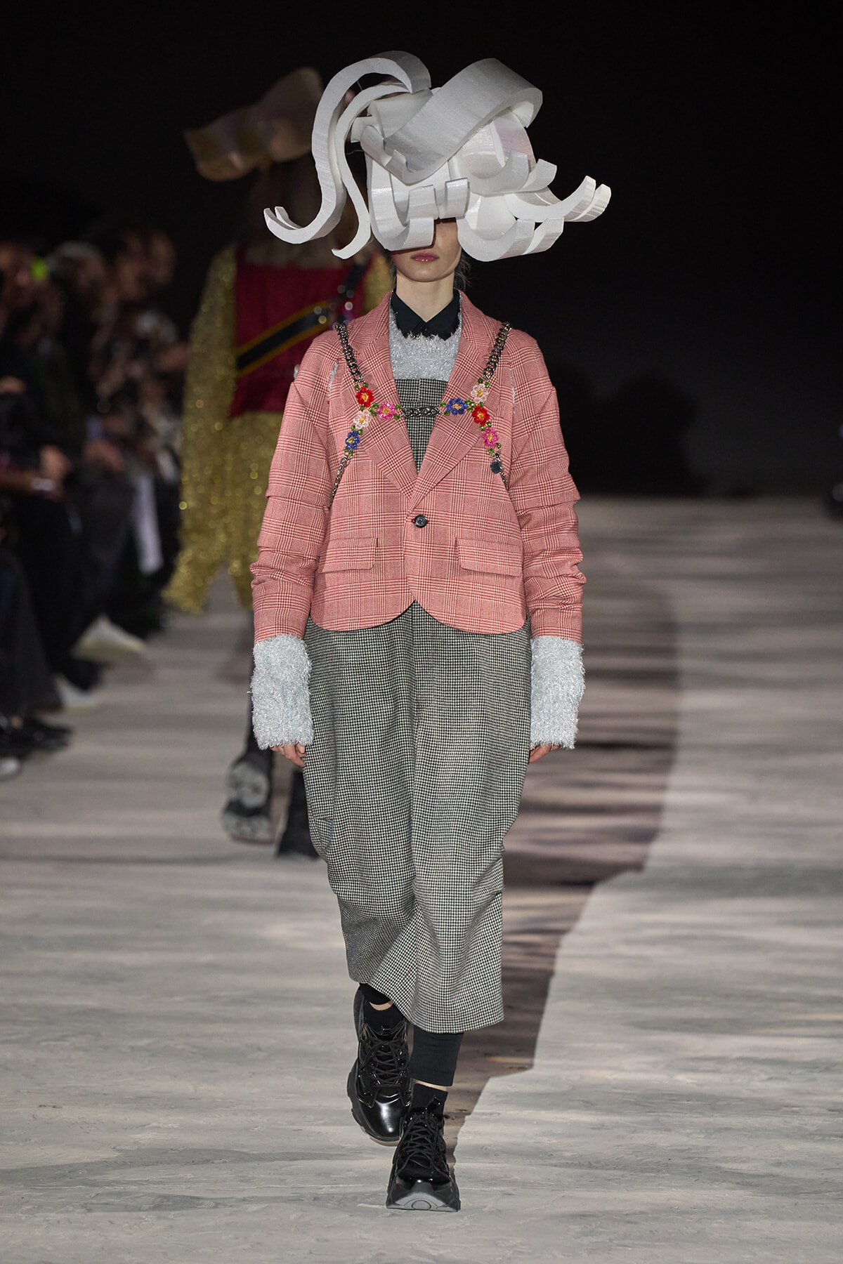 Runway model in pink plaid blazer with floral embroidery over gray check pants and a large white sculptural hat.