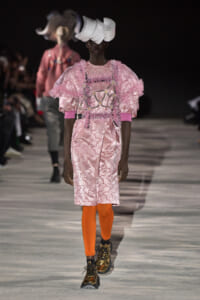 Model walks the runway in a pink embellished dress with sculpted ruffled shoulders, orange tights, metallic boots, and a white sculptural headpiece.