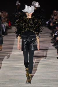 Model on a runway wearing a dramatic black feathered top, black leather shorts, dark tights, gold boots, and a sculptural gray headpiece.