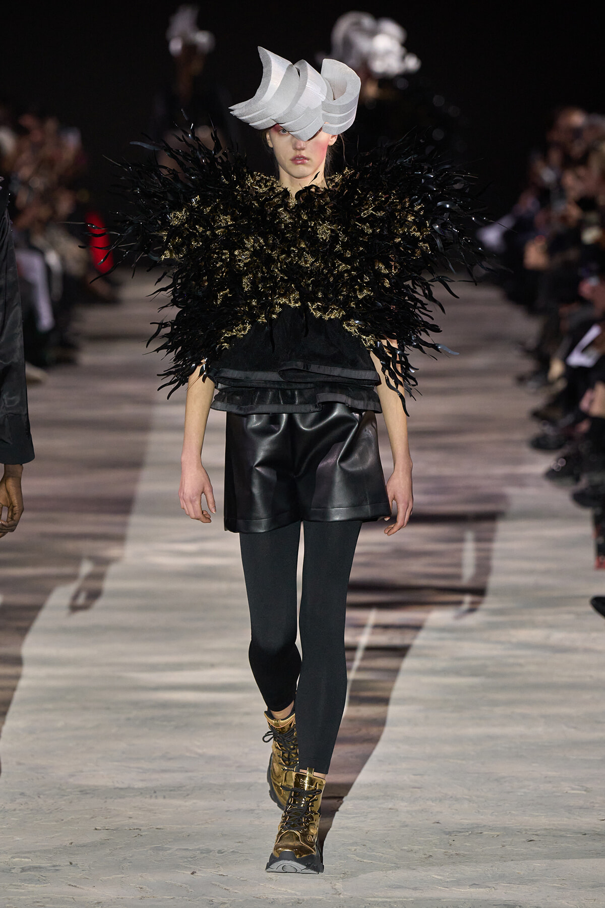 Model on a runway wearing a dramatic black feathered top, black leather shorts, dark tights, gold boots, and a sculptural gray headpiece.