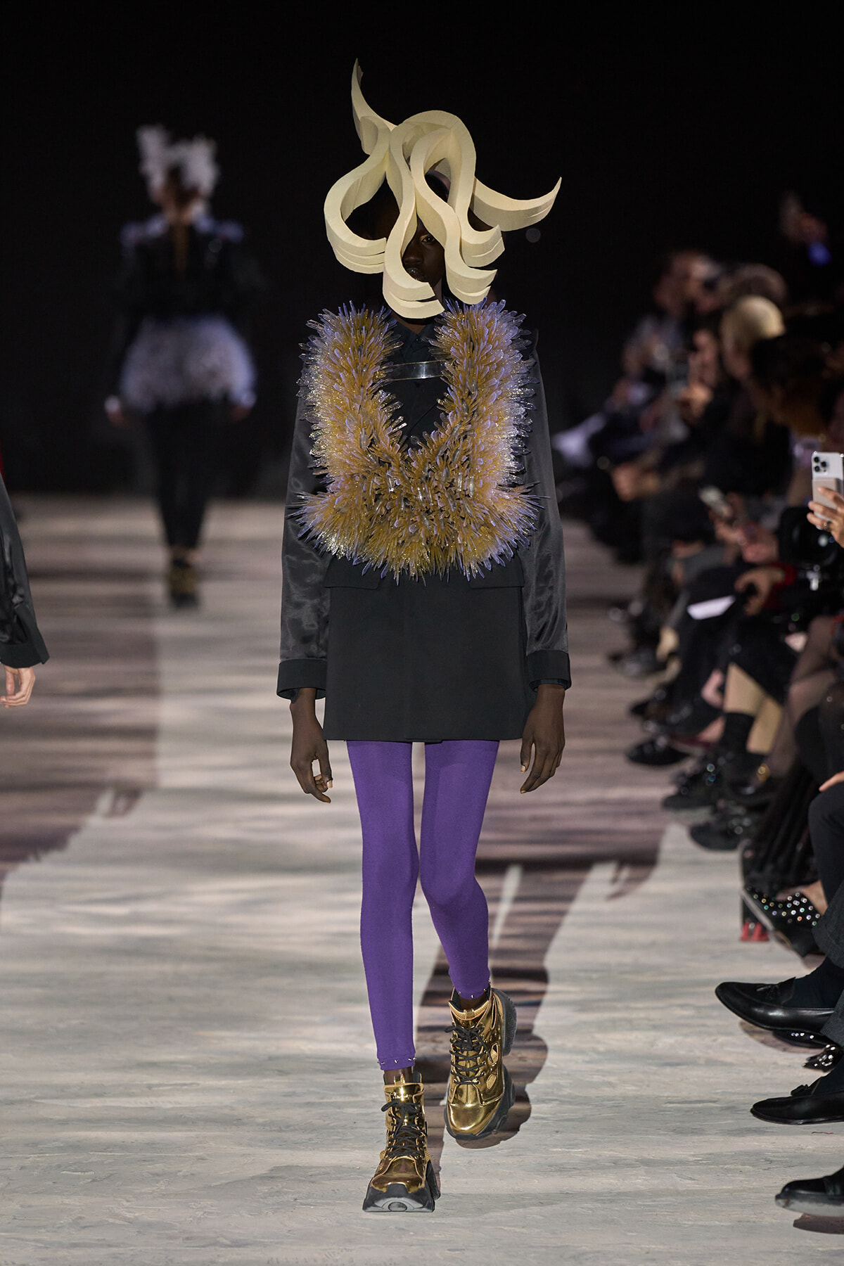 Runway model wearing a large beige sculptural headpiece, gold spiked necklace, black dress, and purple tights.