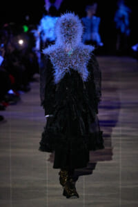 Model on a runway wearing a black textured coat with a blue spiky feather collar covering the head and shoulders, illuminated in blue lighting.