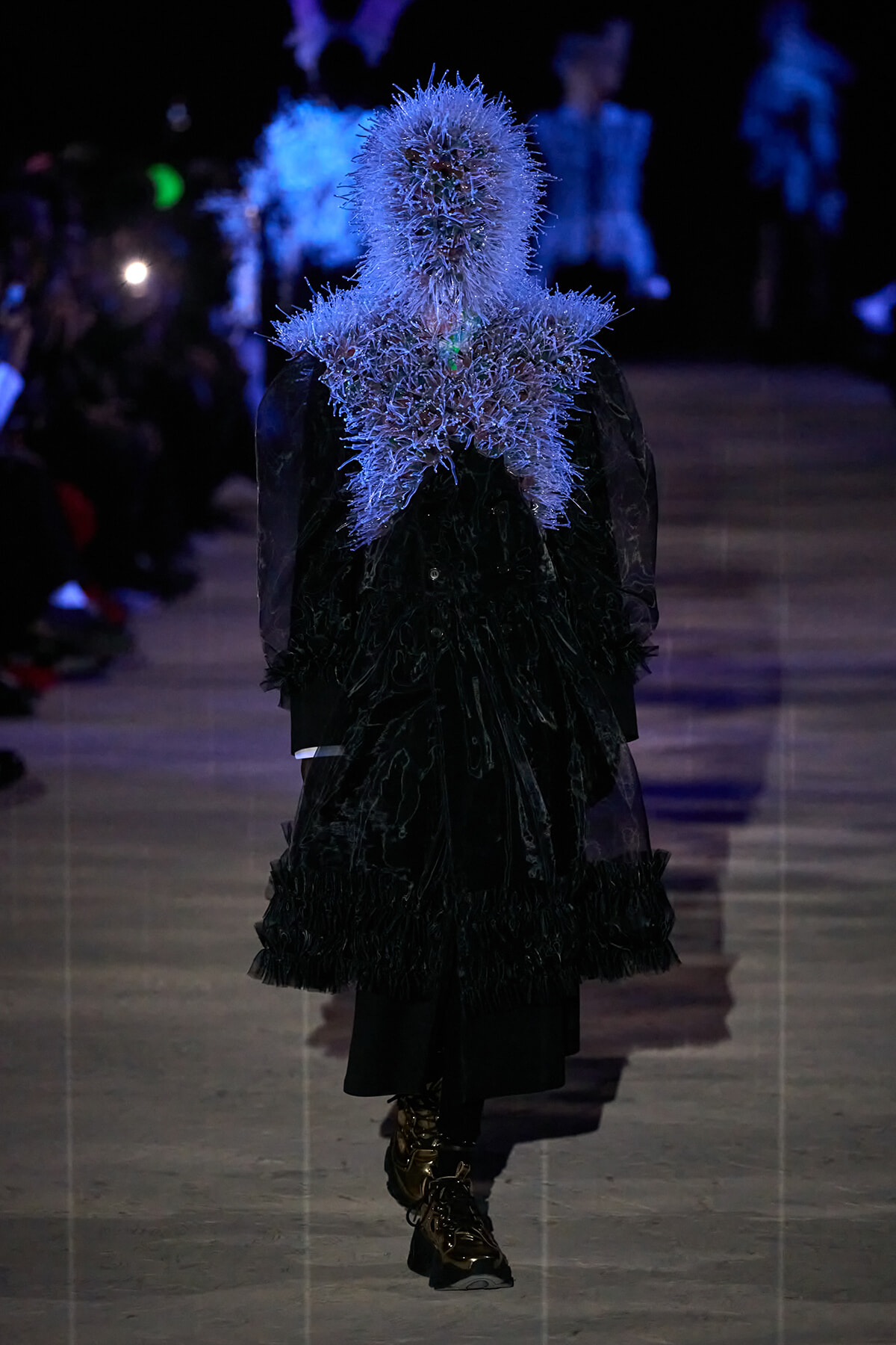 Model on a runway wearing a black textured coat with a blue spiky feather collar covering the head and shoulders, illuminated in blue lighting.