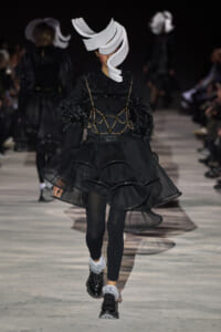 Model on a runway wearing an all-black avant-garde outfit with a white sculptural headpiece and layered sheer skirt.