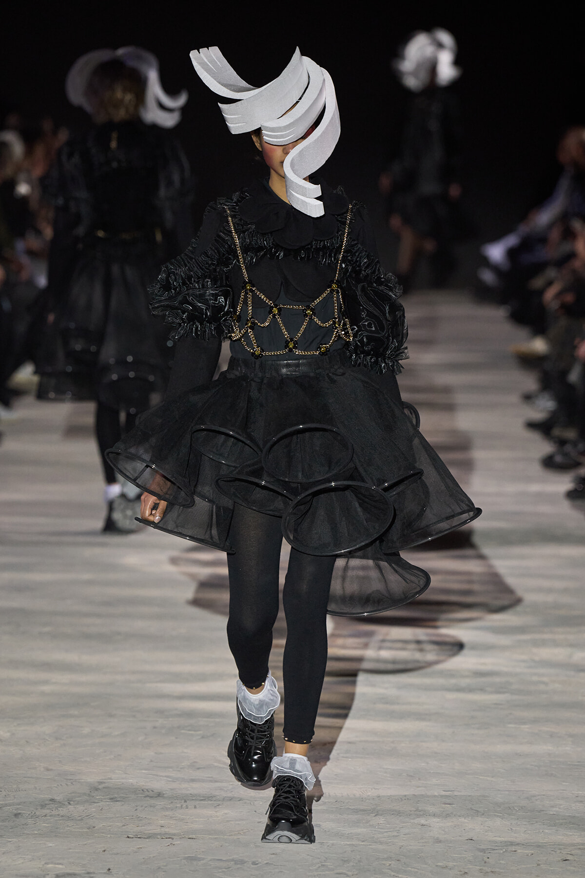 Model on a runway wearing an all-black avant-garde outfit with a white sculptural headpiece and layered sheer skirt.