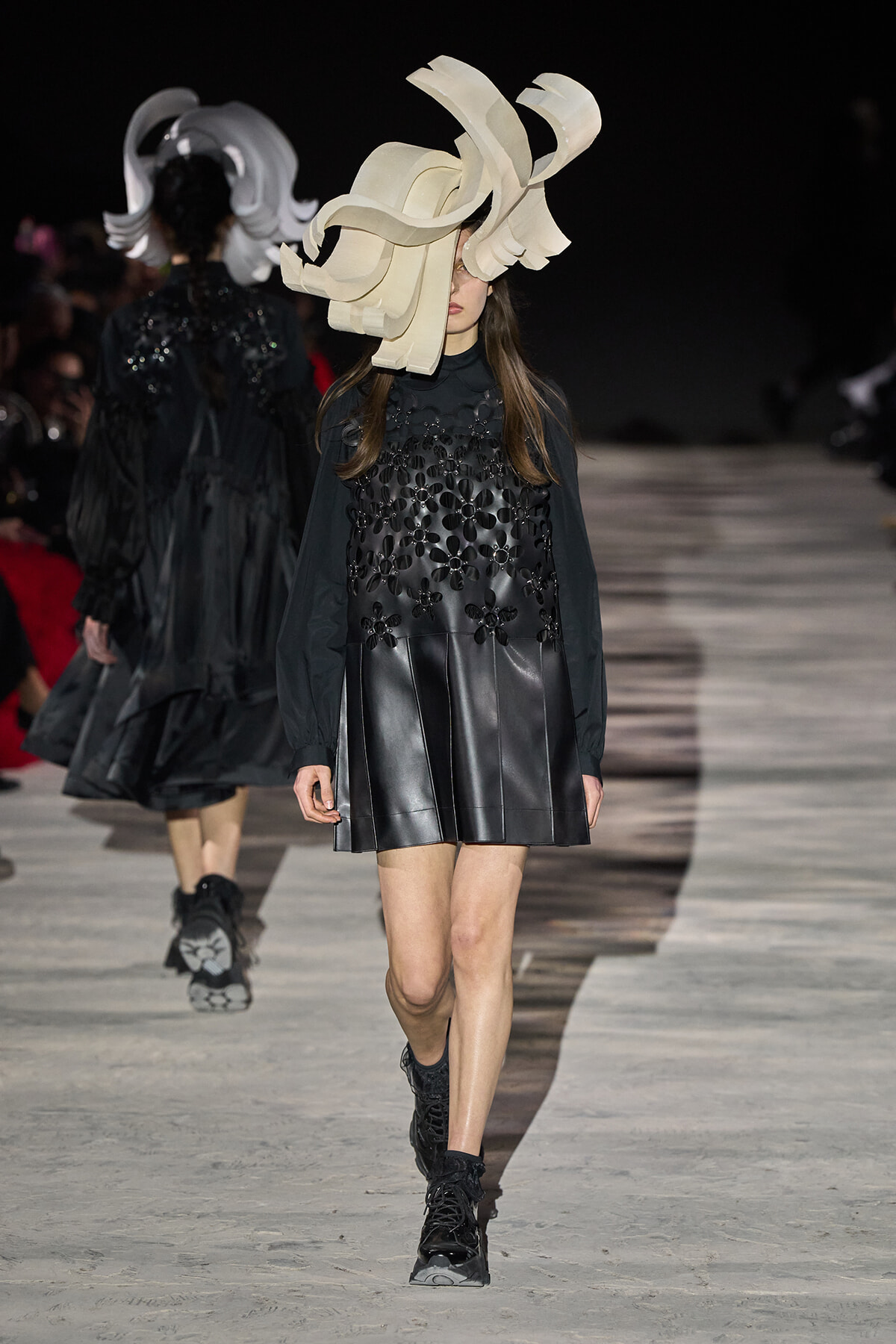 Model walking a runway in a black leather dress with floral appliqués and a large, cream sculptural headpiece with curling ribbons.