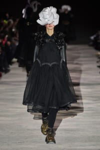 Model on a runway wearing a voluminous black dress with large floral shoulder embellishments and a dramatic white sculptural headpiece, gold shoes visible at the hem.