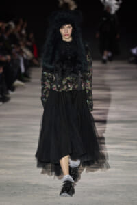 Model on a runway wearing a black floral jacket with an oversized black fur hood and a layered black tulle skirt with boots.