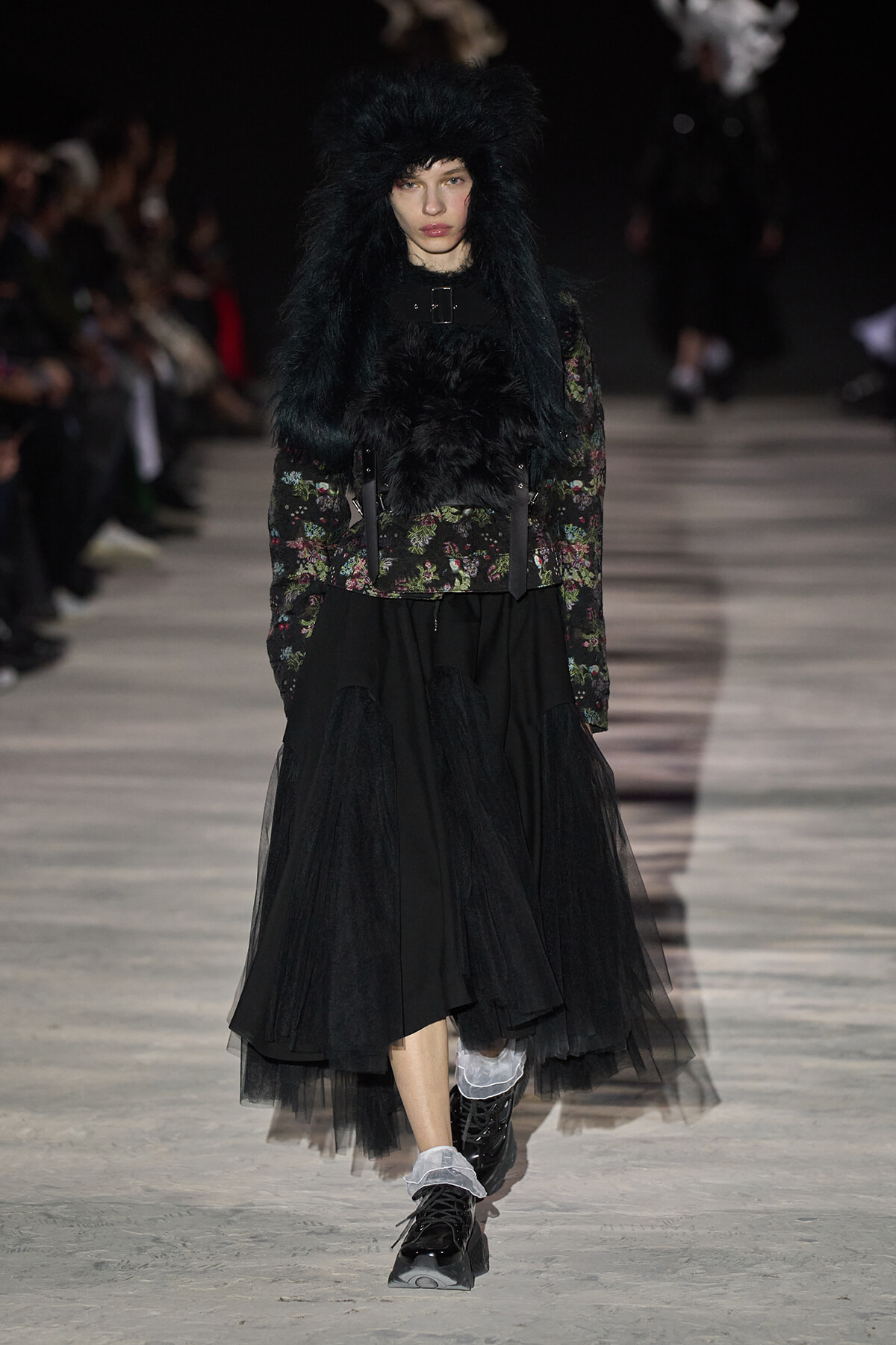 Model on a runway wearing a black floral jacket with an oversized black fur hood and a layered black tulle skirt with boots.