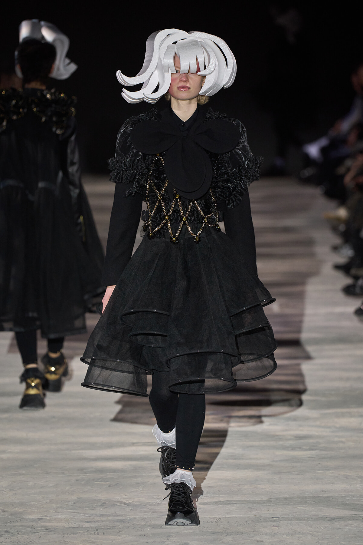 Model on a runway wearing a black multi-tiered tulle dress with a large bow at the neck and a white sculptural headpiece; gold chain lattice belt adds contrast.