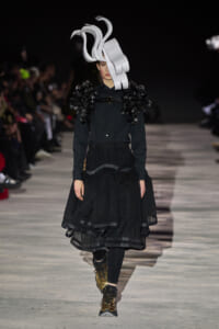Model on a runway wearing an all-black ensemble with oversized sculptural silver headpiece and voluminous layered skirt; gold boots add contrast.