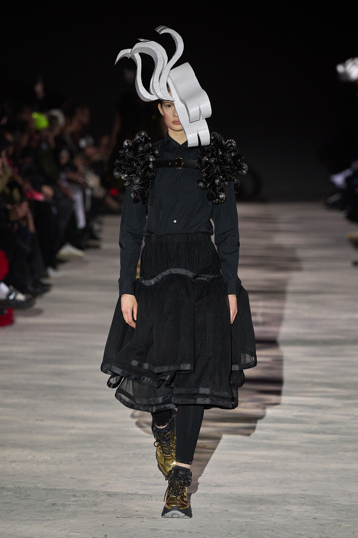 Model on a runway wearing an all-black ensemble with oversized sculptural silver headpiece and voluminous layered skirt; gold boots add contrast.