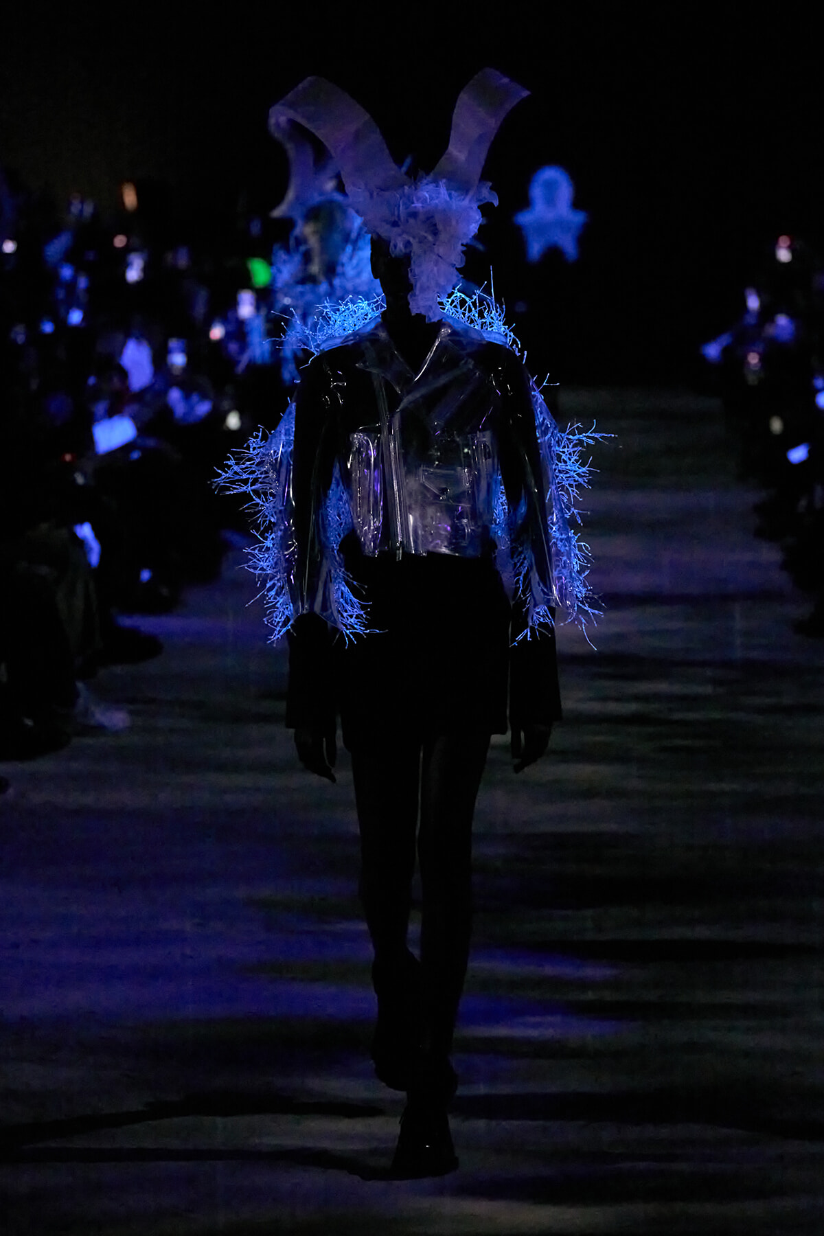 Model on a dark runway wearing an avant-garde outfit with neon blue fringe and a large white bunny-ear headpiece under blue spotlight.