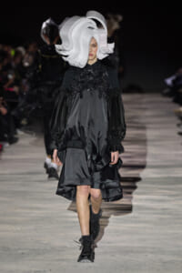 Model walks the runway in a black satin dress with voluminous puff sleeves and a large white sculptural headpiece.