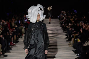Model walks the runway in a dramatic oversized white sculptural hat and a black satin dress with voluminous sleeves, audience on either side observing.