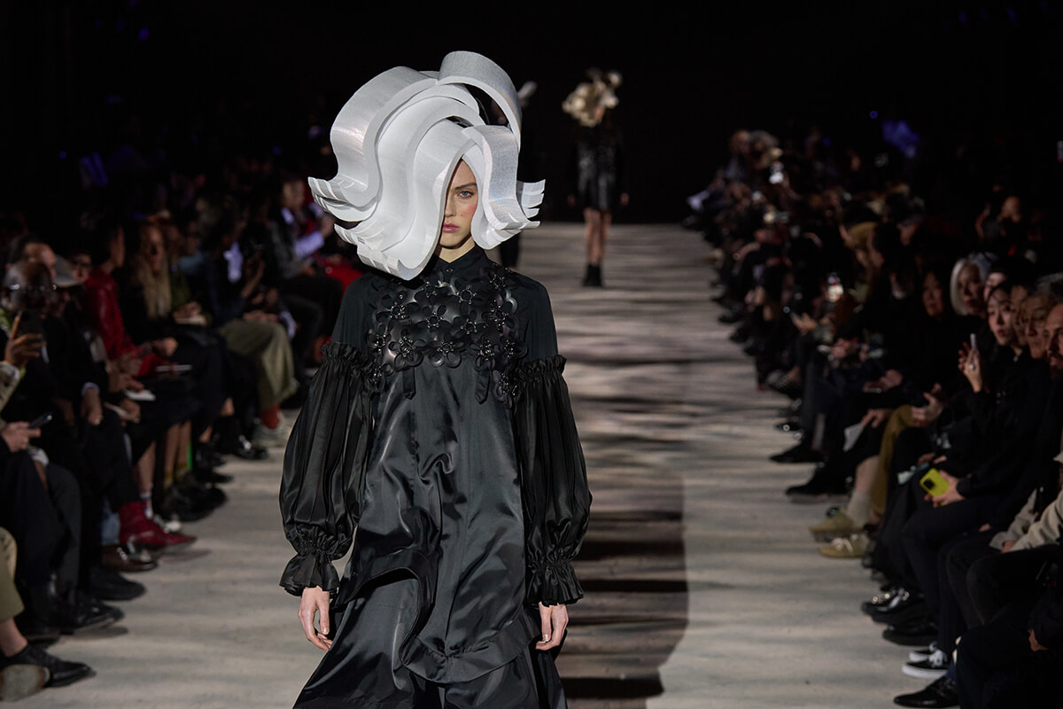 Model walks the runway in a dramatic oversized white sculptural hat and a black satin dress with voluminous sleeves, audience on either side observing.