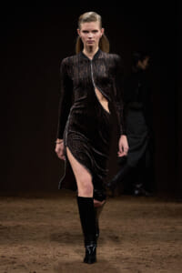 Model walks the runway in a black textured dress with a side cut-out and knee-high boots.