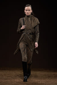 Runway model wearing a brown patterned knit suit with a belted waist and knee-high black boots, walking on a dark catwalk.