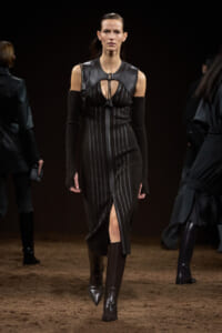 Female model walks down a runway in a black, pin-striped dress with a leather yoke and matching armwarmers, knee-high boots.
