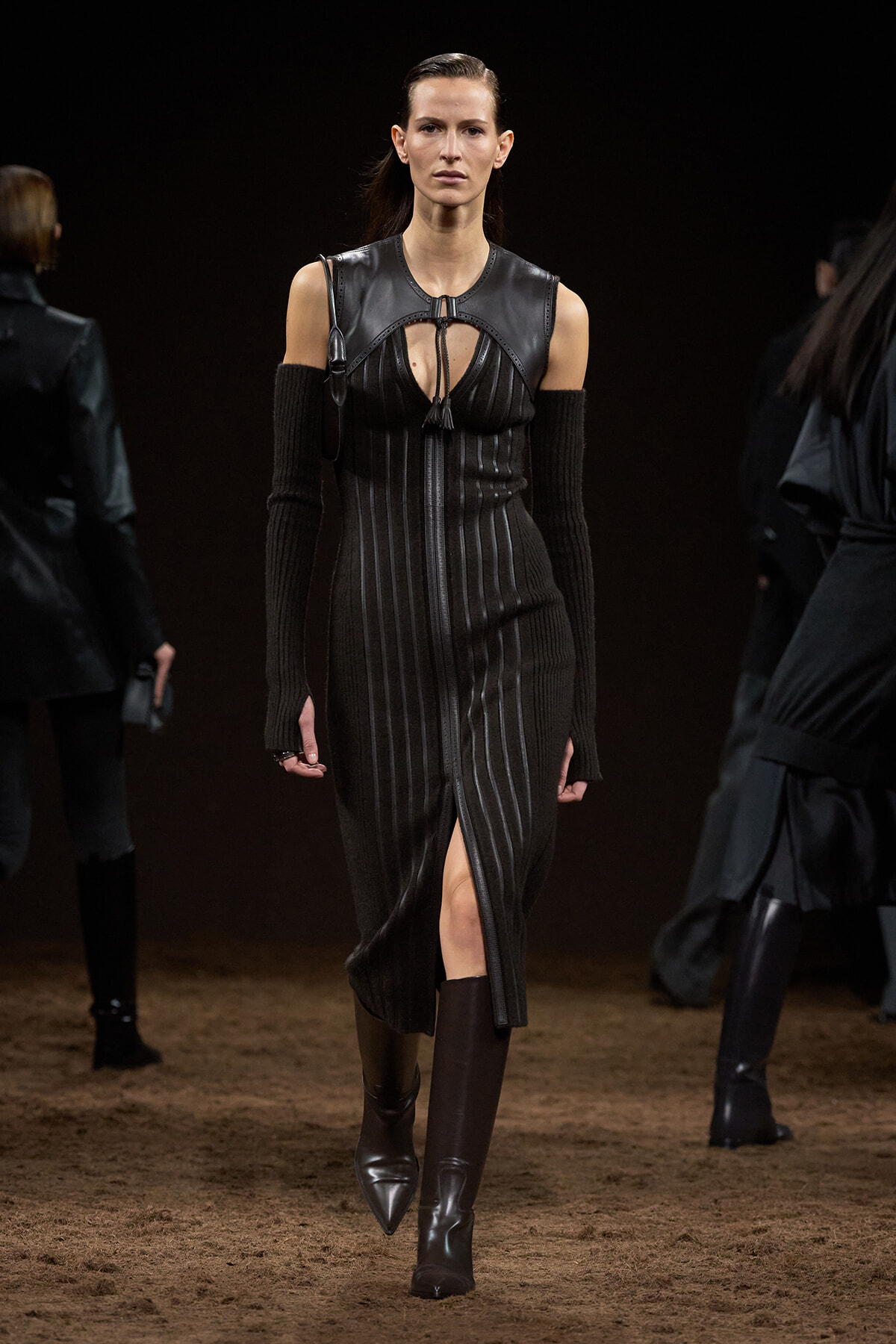 Female model walks down a runway in a black, pin-striped dress with a leather yoke and matching armwarmers, knee-high boots.