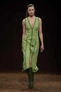 Runway model in a lime-green sleeveless V-neck dress with a waist belt, front slit, and knee-high green boots on a dark backdrop, mid-stride.