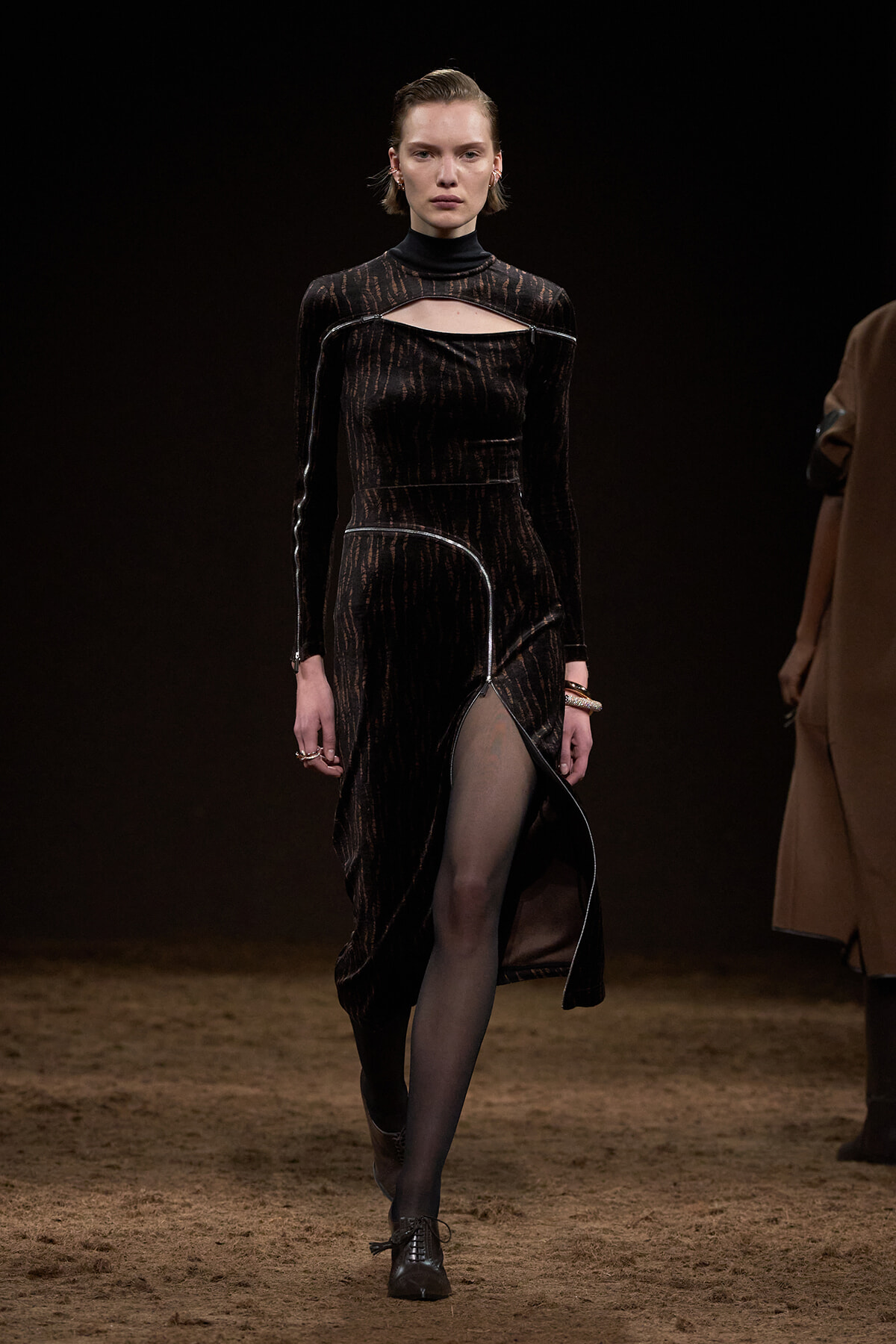 Model on a dark runway wearing a black textured velvet dress with a cutout neckline, zippers, and a high slit, paired with black tights and lace-up boots.