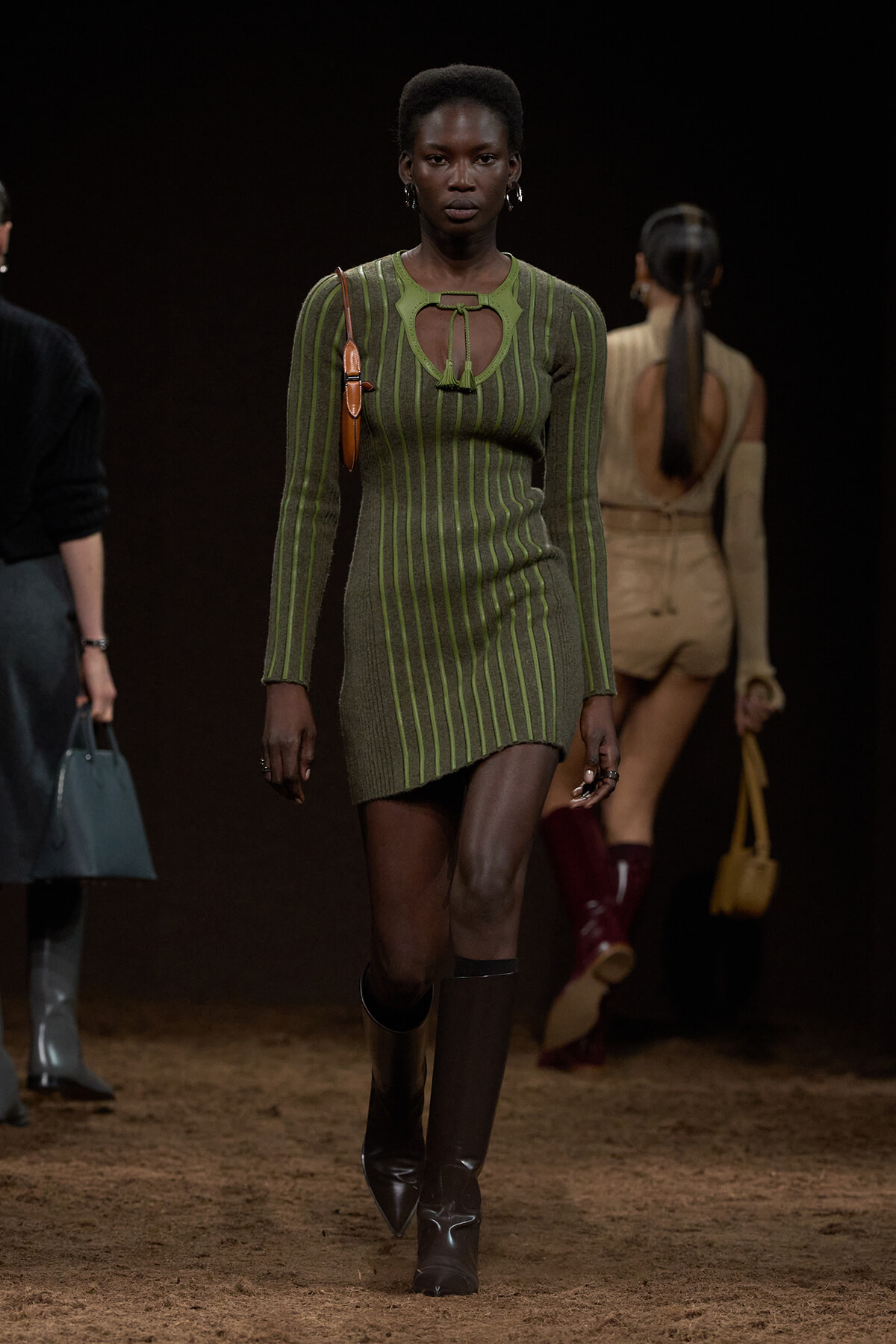 Model walking a runway in a green striped knit mini dress with a cutout neckline, brown knee-high boots, and a small brown shoulder bag.