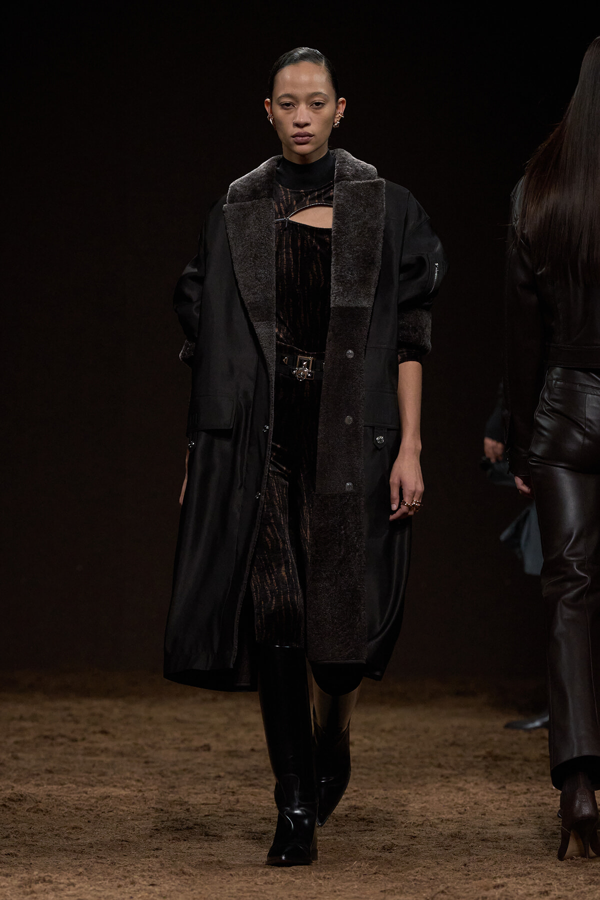 Female model walking a runway in a black coat with shearling trim over a velvet dress and knee-high boots.