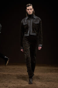 Model walking a fashion runway in a dark velvet jumpsuit with black leather panels and belt, on a brown dirt catwalk.