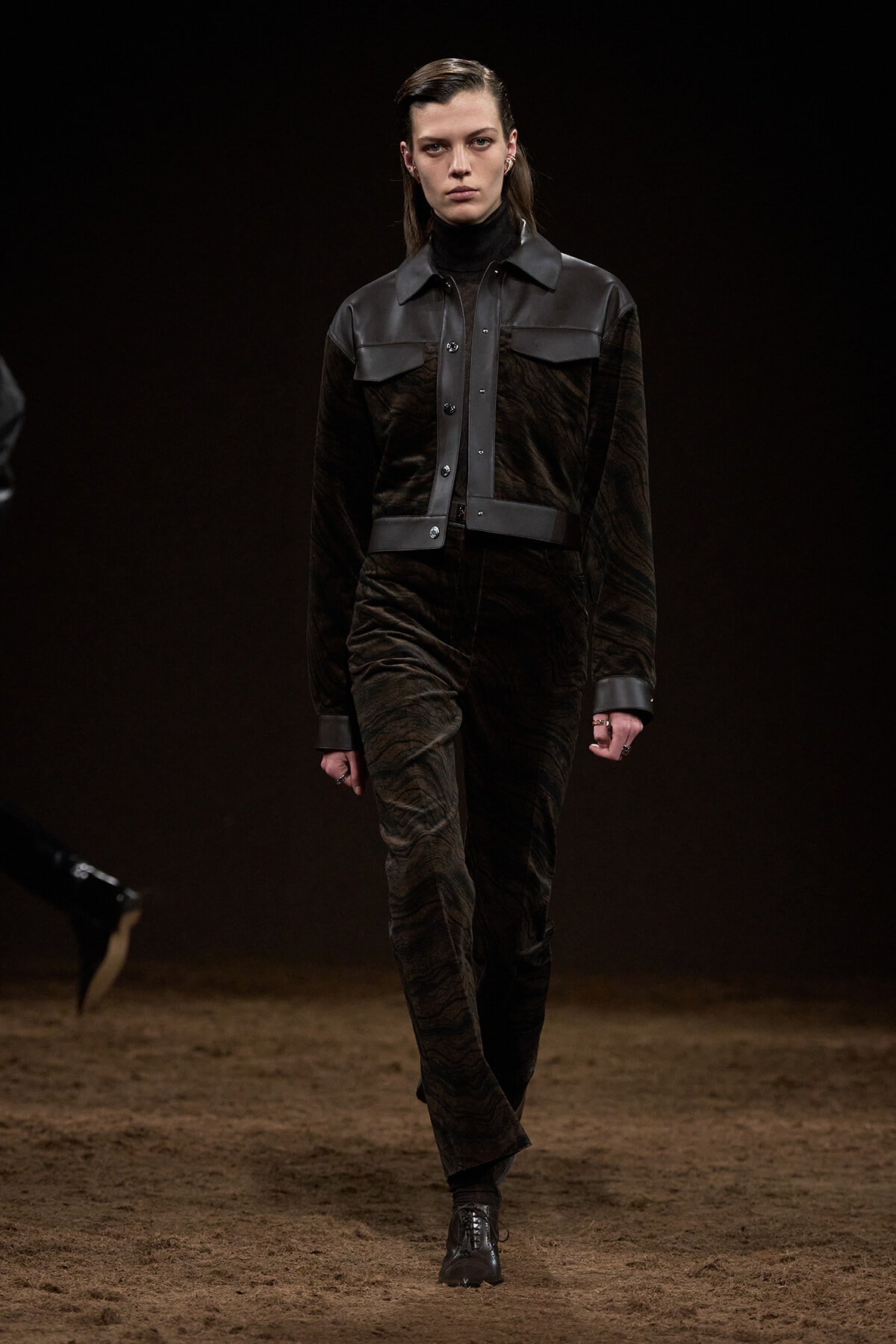Model walking a fashion runway in a dark velvet jumpsuit with black leather panels and belt, on a brown dirt catwalk.