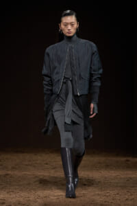 Model walks a dark-toned runway wearing a navy bomber jacket over a layered gray outfit with tall black boots.