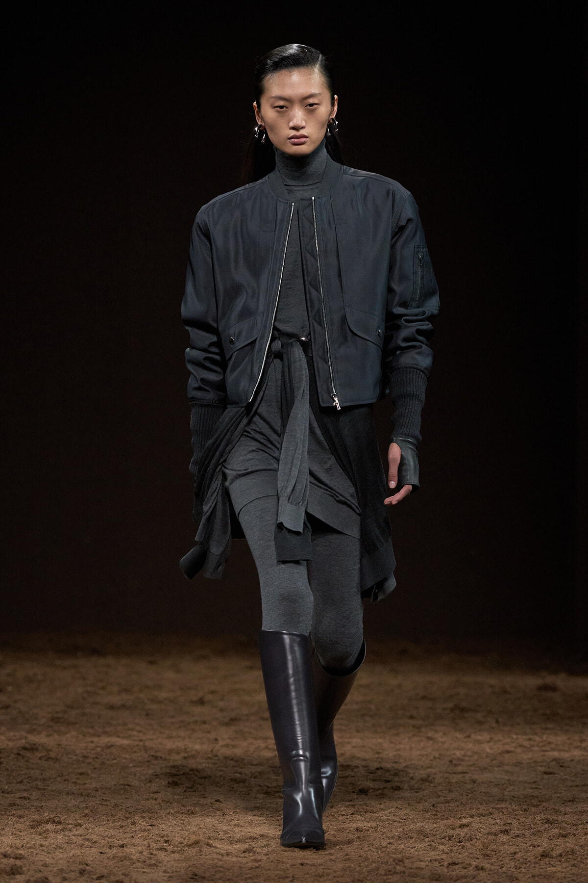 Model walks a dark-toned runway wearing a navy bomber jacket over a layered gray outfit with tall black boots.