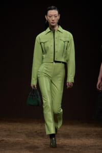Model on a runway wearing a lime-green cropped jacket and matching high-waisted pants, carrying a dark green handbag.