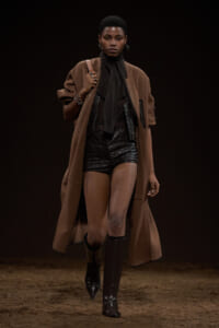 Model walking a runway in a camel-brown long coat over a black quilted outfit with knee-high boots and a black scarf.