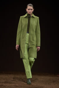 Model on a runway wearing an olive-green suede coat over a matching green suit against a dark backdrop.
