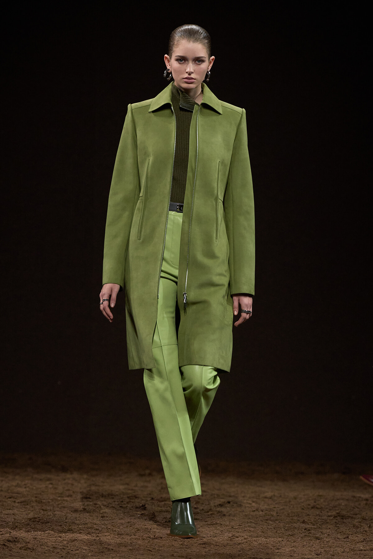 Model on a runway wearing an olive-green suede coat over a matching green suit against a dark backdrop.