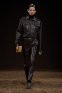 Model walking a fashion show in a black leather cropped jacket and matching pants, holding a small tan clutch.