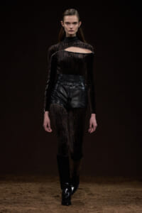 Model on a runway wearing a black sheer patterned bodysuit with a chest cutout, paired with high-waisted black leather shorts and tall black boots.