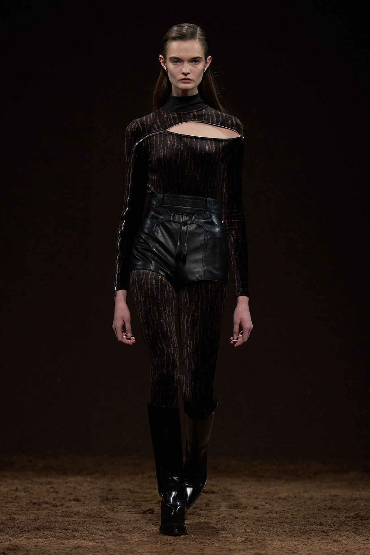 Model on a runway wearing a black sheer patterned bodysuit with a chest cutout, paired with high-waisted black leather shorts and tall black boots.