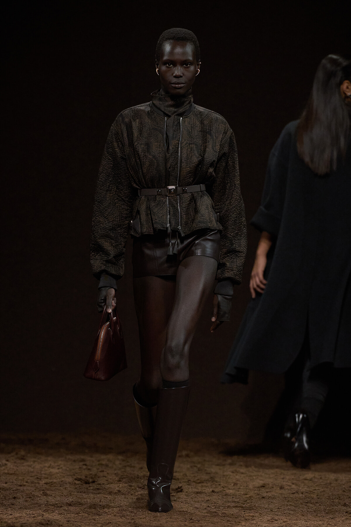 Model walks the runway in a dark textured zip jacket cinched with a belt, black leather shorts, tall boots, and a brown handbag in hand.