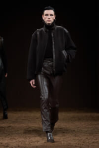Model walking a runway in a black oversized jacket, turtleneck, and dark leather pants.