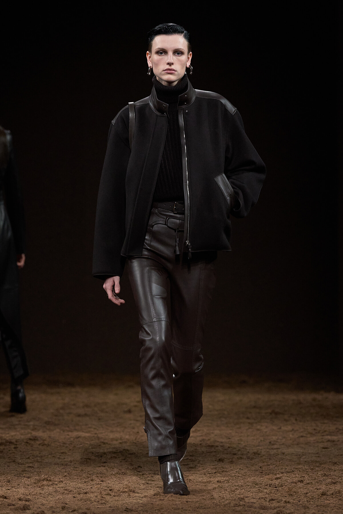Model walking a runway in a black oversized jacket, turtleneck, and dark leather pants.