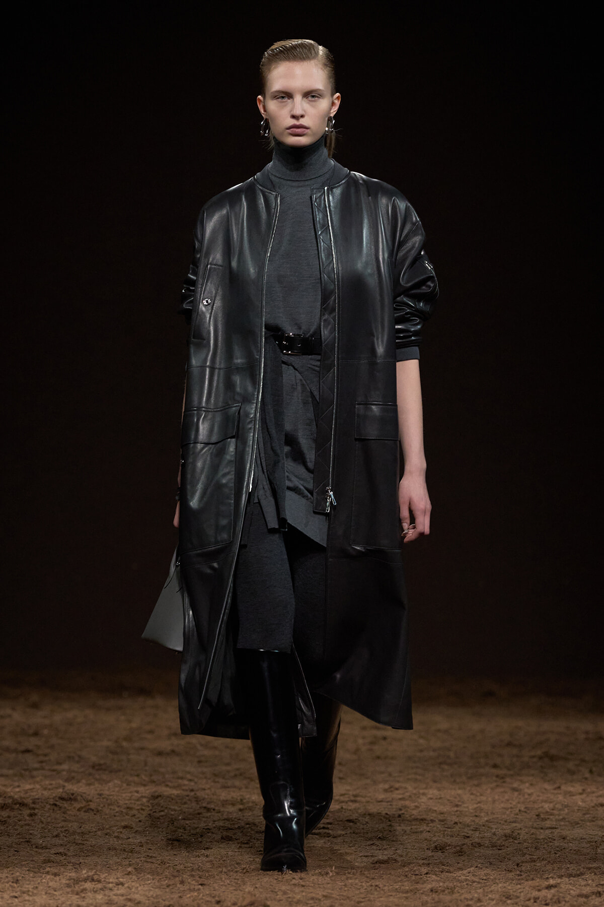 Model walking a runway in a long black leather coat over a dark grey turtleneck and belt, with knee-high black boots.