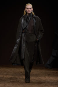 Model on a runway wearing a dark/brown monochrome outfit with a long coat and leather accents.