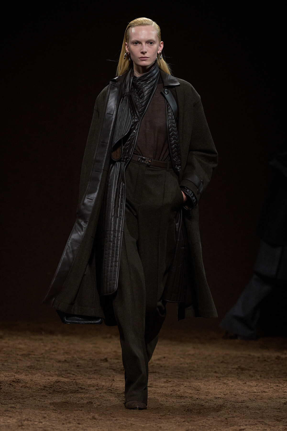 Model on a runway wearing a dark/brown monochrome outfit with a long coat and leather accents.