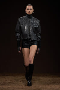 Model walking a fashion runway in a black quilted leather jacket, matching shorts, and knee-high boots against a dark backdrop.