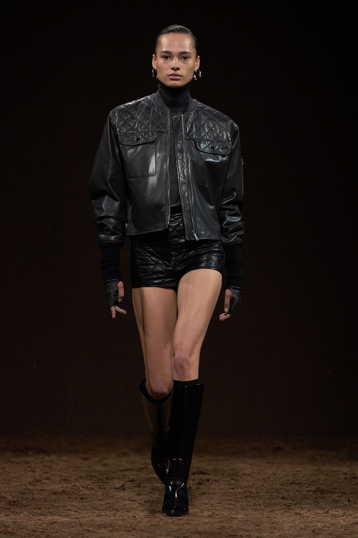 Model walking a fashion runway in a black quilted leather jacket, matching shorts, and knee-high boots against a dark backdrop.
