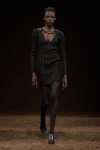 Model walking a runway in a black pinstripe knit dress with a cutout neckline and tie detail at the chest.