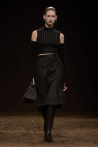 Runway model in an all-black outfit: cold-shoulder knit top, lace-up waist skirt, knee-high boots, and a small handbag.